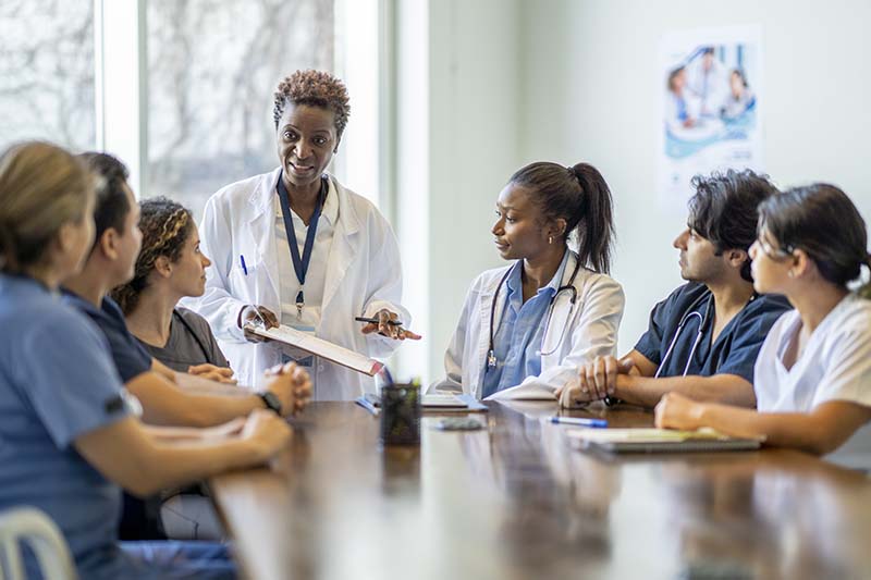 A diverse team of nurses gathers around a table for a meeting, with one standing and leading the discussion, illustrating collaboration and engagement in a clinical setting.