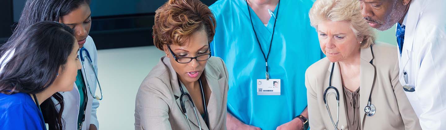 A group of healthcare professionals, including nurse administrators, are gathered around a table reviewing charts and graphs. There is a mix of attentiveness and discussion among the group, with some individuals pointing towards the documents. The setting suggests a collaborative meeting in a medical office environment.