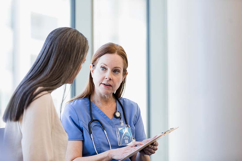 A nurse in blue scrubs with a stethoscope around her neck is attentively listening to a patient while holding a clipboard. They are standing in a brightly lit room with a large window in the background.