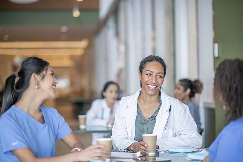 A group of nurses are in a medical center cafeteria seated at a table. They are smiling and laughing and engaged in a pleasant conversation.
