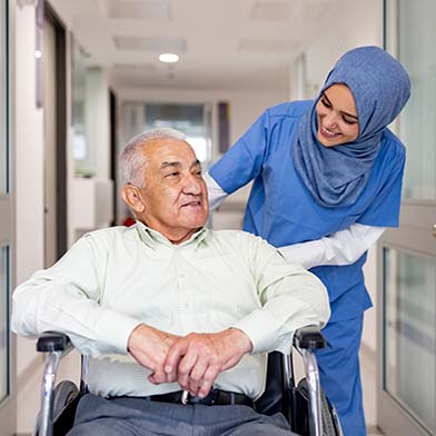 A nurse in blue scrubs and a hijab is gently pushing an elderly gentleman in a wheelchair through a hospital corridor. The nurse is leaning forward slightly, smiling and engaging with the patient, who looks relaxed and content.