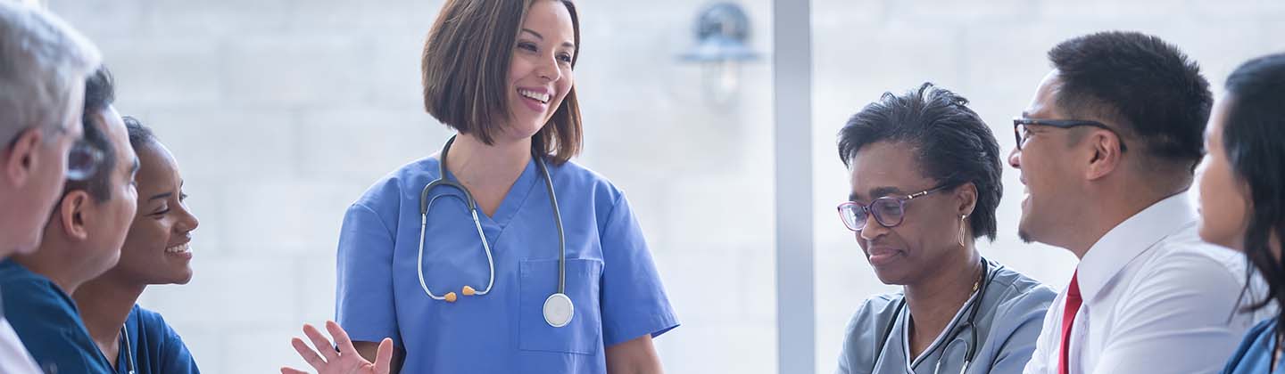 A young female nurse is wearing blue scrubs and is standing at the head of a conference table in front of a group of seated health care workers. She is smiling and appears to be sharing a story.