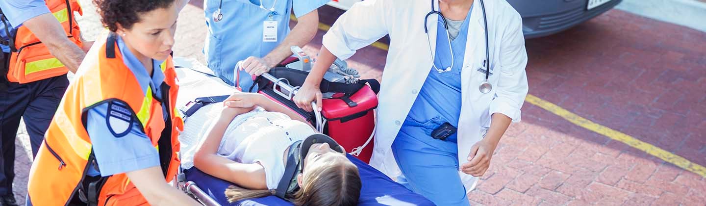 Two Emergency Room staff members are helping a team of emergency medical technicians to push a stretcher into the Emergency Department. A young child wearing a neck brace is calmly laying on the stretcher.