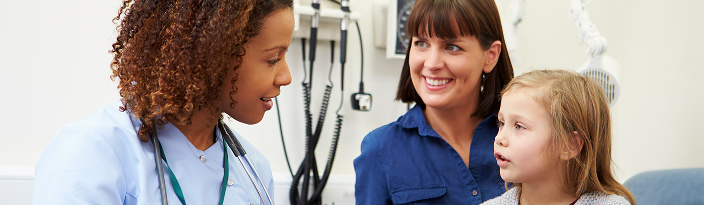 A nurse wearing light blue scrubs is speaking with a mother and her young daughter during a clinic appointment. The nurse is holding a folder and explaining something to the young girl, and the mother is smiling.