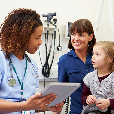 A nurse wearing light blue scrubs is speaking with a mother and her young daughter during a clinic appointment. The nurse is holding a folder and explaining something to the young girl, and the mother is smiling.